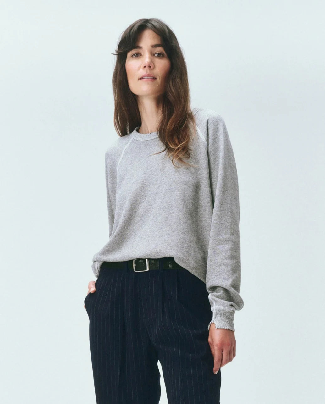A woman with long brown hair models The Great Inc.'s The College Sweatshirt VARSITY GREY and dark pinstripe pants, standing confidently against a plain light background.
