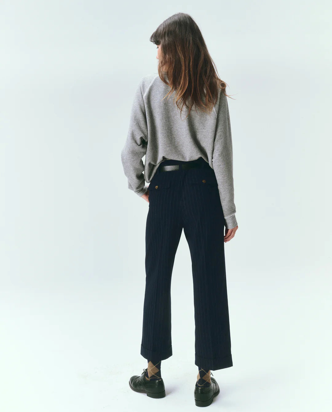 A person with long brown hair stands facing away, wearing The Great Inc.'s The College Sweatshirt VARSITY GREY, dark pinstripe pants, and black shoes against a plain light background.