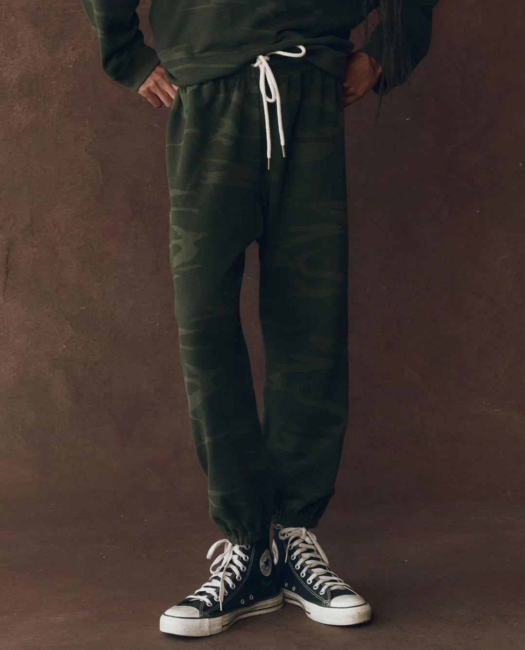 Wearing The Great Inc.&#39;s The Stadium Sweatpant in dark green camouflage with a white drawstring and black high-top Converse sneakers, a person stands against a brown textured background; upper body not visible, evoking vintage athletic style.
