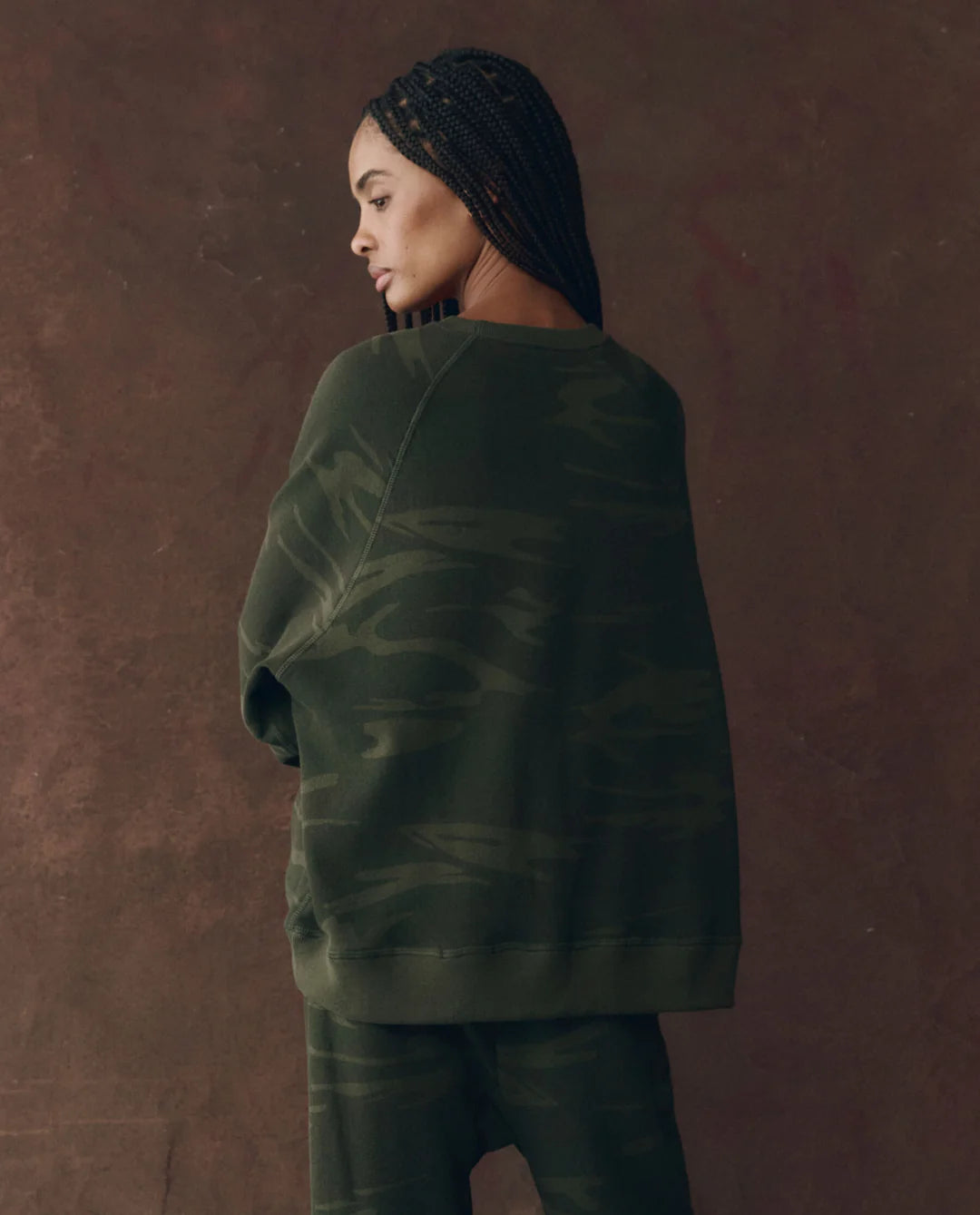 A woman with braided hair wears The Great Inc.'s dark green camo-patterned The Slouch Sweatshirt and matching pants, both made in Los Angeles, as she stands slightly turned to the side against a brown background.