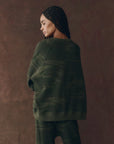 A woman with braided hair wears The Great Inc.'s dark green camo-patterned The Slouch Sweatshirt and matching pants, both made in Los Angeles, as she stands slightly turned to the side against a brown background.