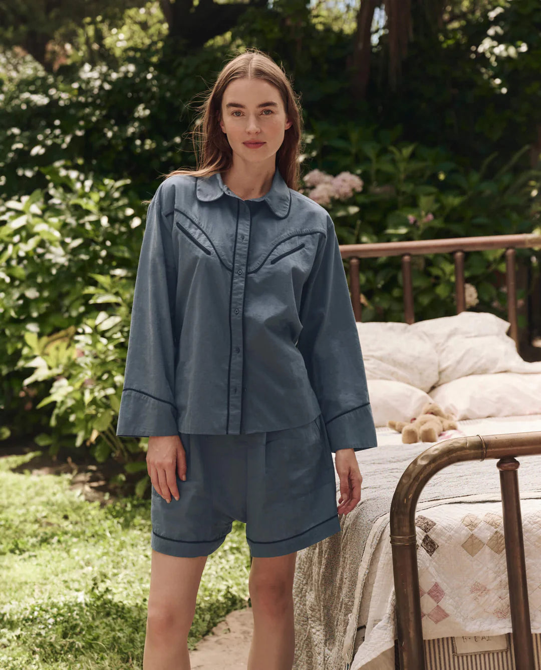A woman poses outdoors beside a brass bed, wearing The Great Inc.'s The Pajama Shirt and matching shorts. Surrounded by greenery and flowers, she exudes relaxed nostalgia in this natural setting.