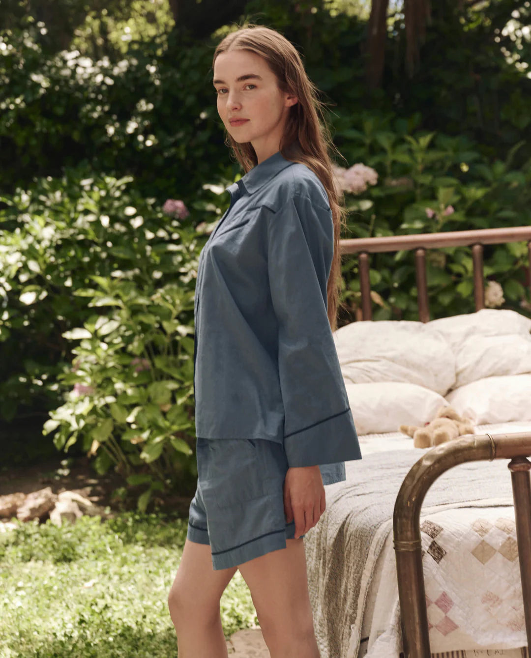 A woman with long hair stands outdoors by a vintage brass bed with white bedding, wearing The Pajama Shirt from The Great Inc. Lush green plants and flowers in the background create a dreamy atmosphere.