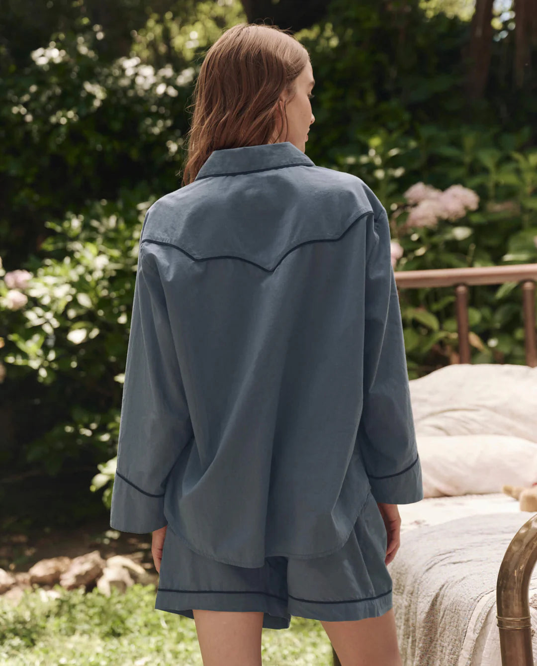 A person with long, light brown hair stands outdoors, back to camera, wearing The Pajama Shirt by The Great Inc. in blue with matching shorts. Greenery and a bed with pale bedding add vintage charm to the scene.