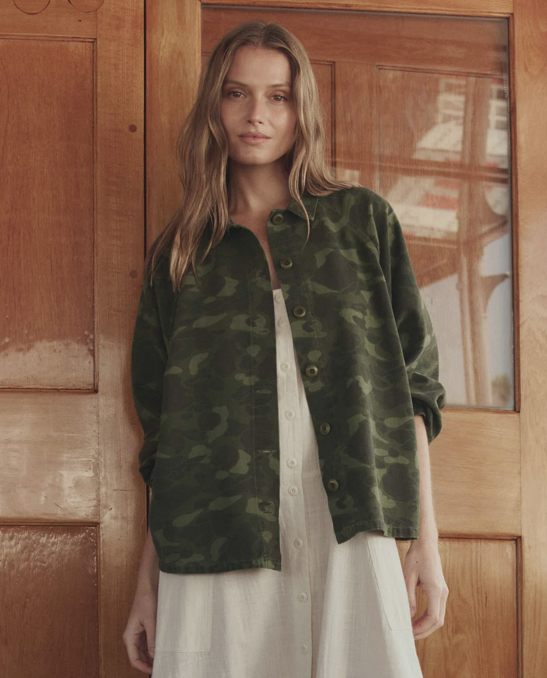 A woman with long, wavy hair stands indoors by wooden double doors, wearing The Great Inc.'s green 1940s camo cotton sateen Commodore Jacket over a white dress; a glass window reflects the outdoors behind her.