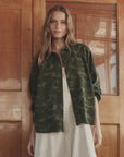 A woman with long, wavy hair stands indoors by wooden double doors, wearing The Great Inc.'s green 1940s camo cotton sateen Commodore Jacket over a white dress; a glass window reflects the outdoors behind her.
