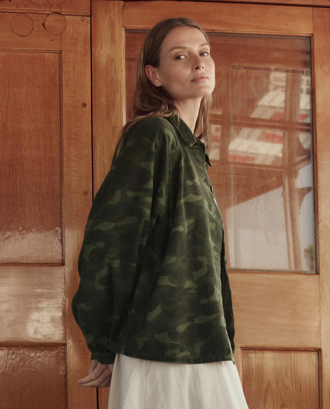 A woman with long hair wears The Great Inc.’s The Commodore Jacket, a vintage-inspired green camouflage piece, over a white dress as she stands before wooden doors, glancing back over her shoulder.