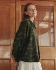 A woman with long hair wears The Great Inc.’s The Commodore Jacket, a vintage-inspired green camouflage piece, over a white dress as she stands before wooden doors, glancing back over her shoulder.