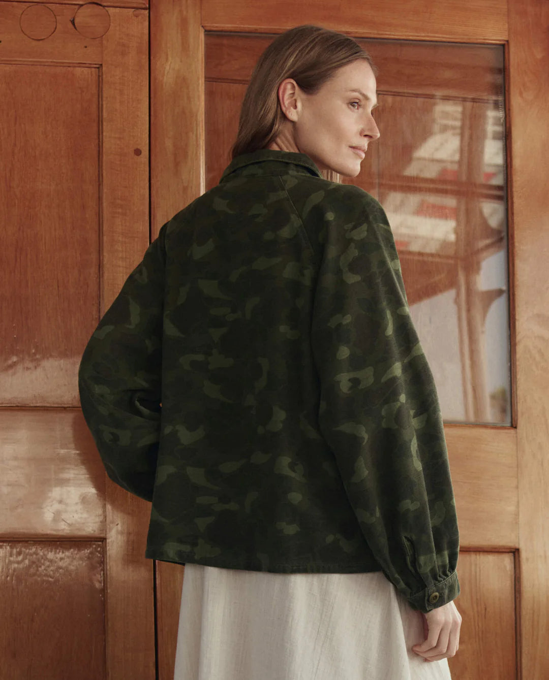 A woman with light brown hair stands indoors, back turned, wearing The Commodore Jacket by The Great Inc. over a light dress in front of wooden doors with glass panes.