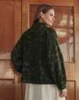 A woman with light brown hair stands indoors, back turned, wearing The Commodore Jacket by The Great Inc. over a light dress in front of wooden doors with glass panes.