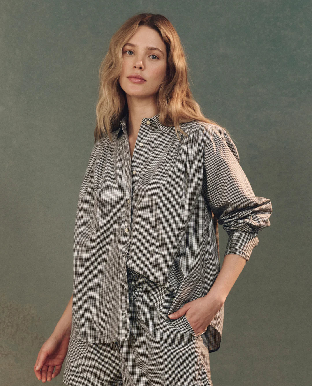 A woman with long blond hair, wearing The Society Top by The Great Inc. as part of a gray and white gingham set, stands against a textured green background, gazing calmly at the camera with one hand in her pocket.