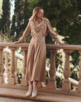 A person stands on a stone terrace, wearing The Great Inc.'s beige "The Western Shirt Woven" blouse with contrast piping and a matching long skirt. They lean against a balustrade surrounded by trees. Beige ankle boots complete their outfit, enhancing the rustic setting.