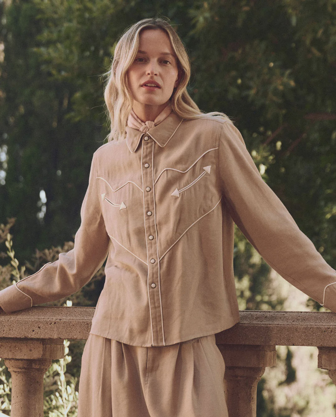 A person with long hair stands outdoors wearing The Great Inc.'s The Western Shirt Woven in tan, paired with a matching skirt. They lean on a stone railing surrounded by trees. The shirt's decorative stitching and contrast piping add distinct flair.