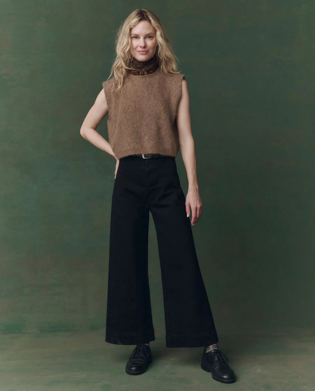 A woman stands against a green background, wearing The Brushed Melange Sweater by The Great Inc. over a patterned turtleneck, paired with wide-leg black pants and black shoes. Her blonde, wavy hair and relaxed posture complete the confident look.