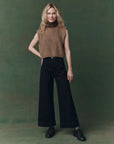 A woman stands against a green background, wearing The Brushed Melange Sweater by The Great Inc. over a patterned turtleneck, paired with wide-leg black pants and black shoes. Her blonde, wavy hair and relaxed posture complete the confident look.