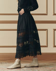 A woman in The Great Inc. Rosette Crochet Canary Skirt with a smocked waistband, a black sweater with decorative buttons, and beige lace-up ankle boots stands on a patterned rug before a tan paneled wall.