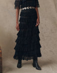 Model wears The Great Inc. "The Winter Mesh Flounce Skirt" in black with a wide silver concho belt and black pointed-toe boots, standing against a neutral textured background; only body from shoulders down is shown.