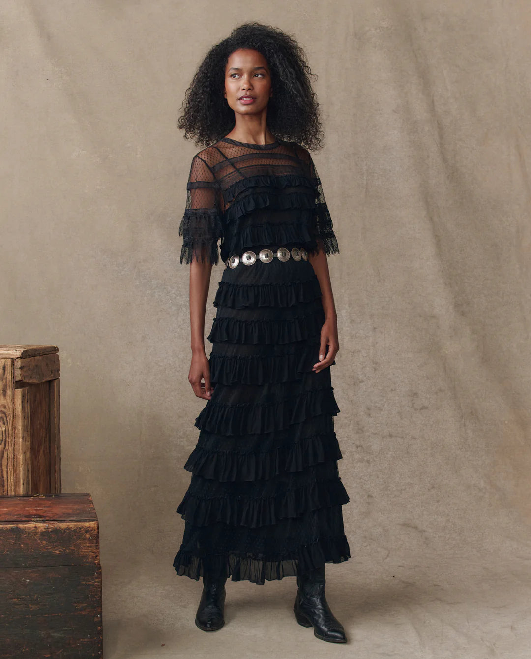 A woman poses before a beige backdrop wearing The Winter Mesh Flounce Skirt by The Great Inc., styled with a black dress featuring tiered ruffles, sheer sleeves, a wide silver belt, black boots, and stacked wooden crates beside her.