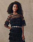 A woman with curly hair models The Great Inc.’s Winter Mesh Flounce Top, featuring sheer dotted mesh, georgette ruffles, and lace accents, styled with a wide metallic belt against a neutral textured backdrop.