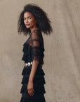 A woman with curly hair models The Great Inc.'s Winter Mesh Flounce Top, a vintage-inspired black piece with georgette ruffles and sheer dotted mesh sleeves, styled with a wide silver belt against a textured neutral backdrop.