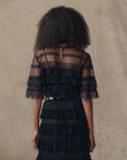 A woman with curly hair faces away, wearing The Great Inc.’s Winter Mesh Flounce Top—a vintage-inspired sheer black piece with dotted mesh and layered georgette ruffles—set against a neutral, textured background.