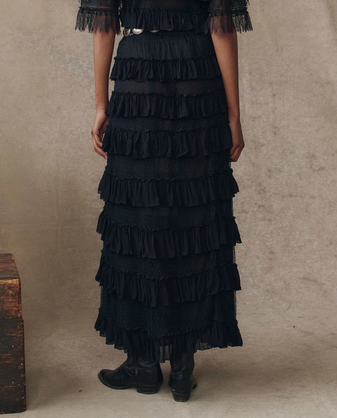 Only the lower body is visible as someone stands against a neutral, textured backdrop, wearing The Great Inc.'s The Winter Mesh Flounce Skirt— a long, black, mesh number with tiered ruffles—paired with black boots.