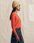 A woman in profile wears The Cashmere Tiny V-Neck Cardigan by The Great Inc., a yellow knit beanie, and a black skirt with white polka dots, standing against a neutral fabric backdrop.