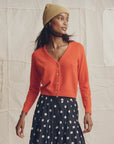 A woman wearing The Great Inc. Cashmere Tiny V-Neck Cardigan in orange, a tan beanie, and a black skirt with white polka dots stands before a beige fabric backdrop, gazing to the side.