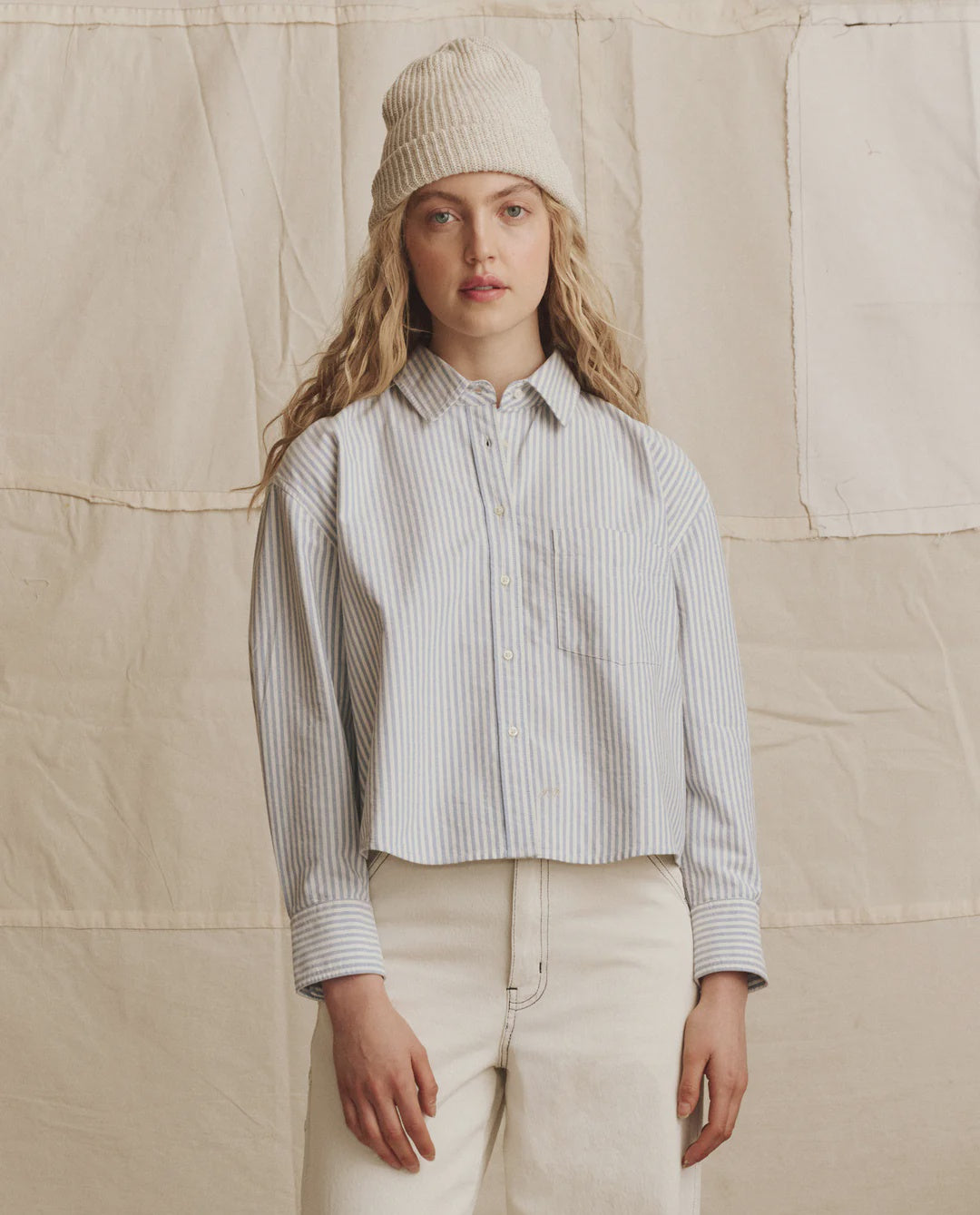 A person with long, wavy blonde hair wears The Cropped Oxford by The Great Inc., a light blue and white striped cotton shirt with a boxy silhouette, paired with a cream knit beanie and light pants, against a beige fabric backdrop.