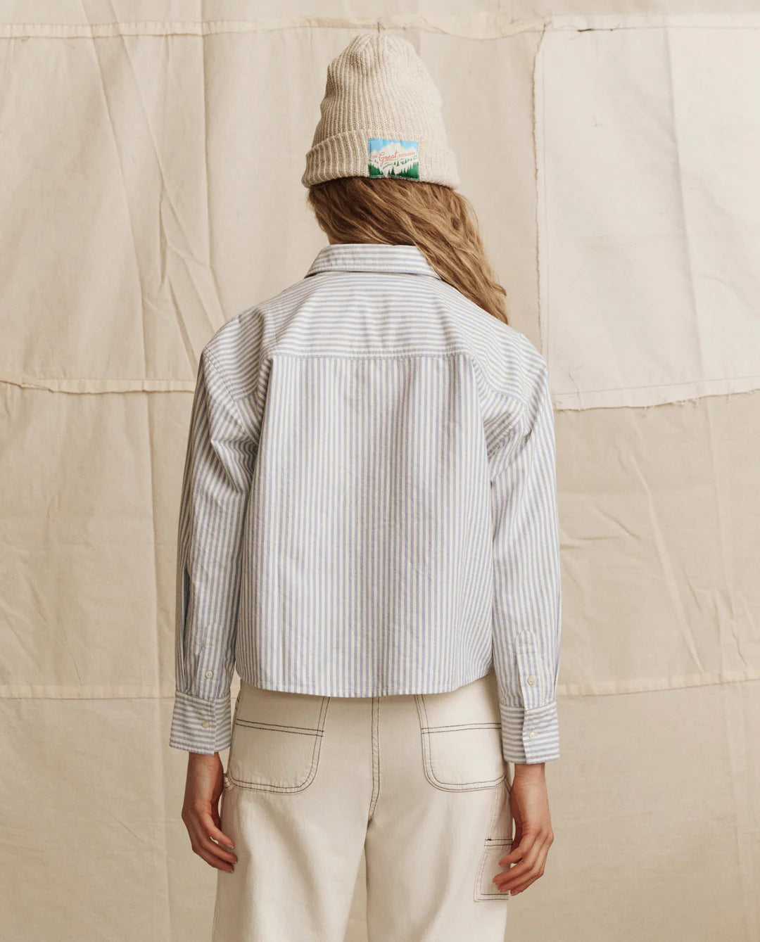 A person stands facing away, wearing The Cropped Oxford by The Great Inc.—a light blue and white striped button-up shirt—paired with a cream knit beanie and high-waisted off-white pants, set against a beige fabric backdrop.