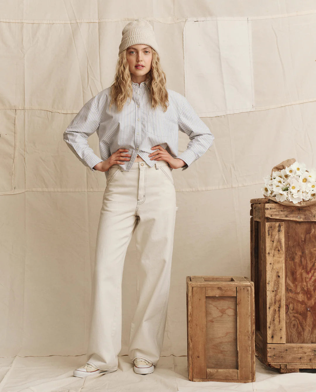 A woman wears The Cropped Oxford by The Great Inc., paired with cream wide-leg pants and a knit beanie, standing indoors beside stacked wooden crates with white flowers against a neutral fabric backdrop.