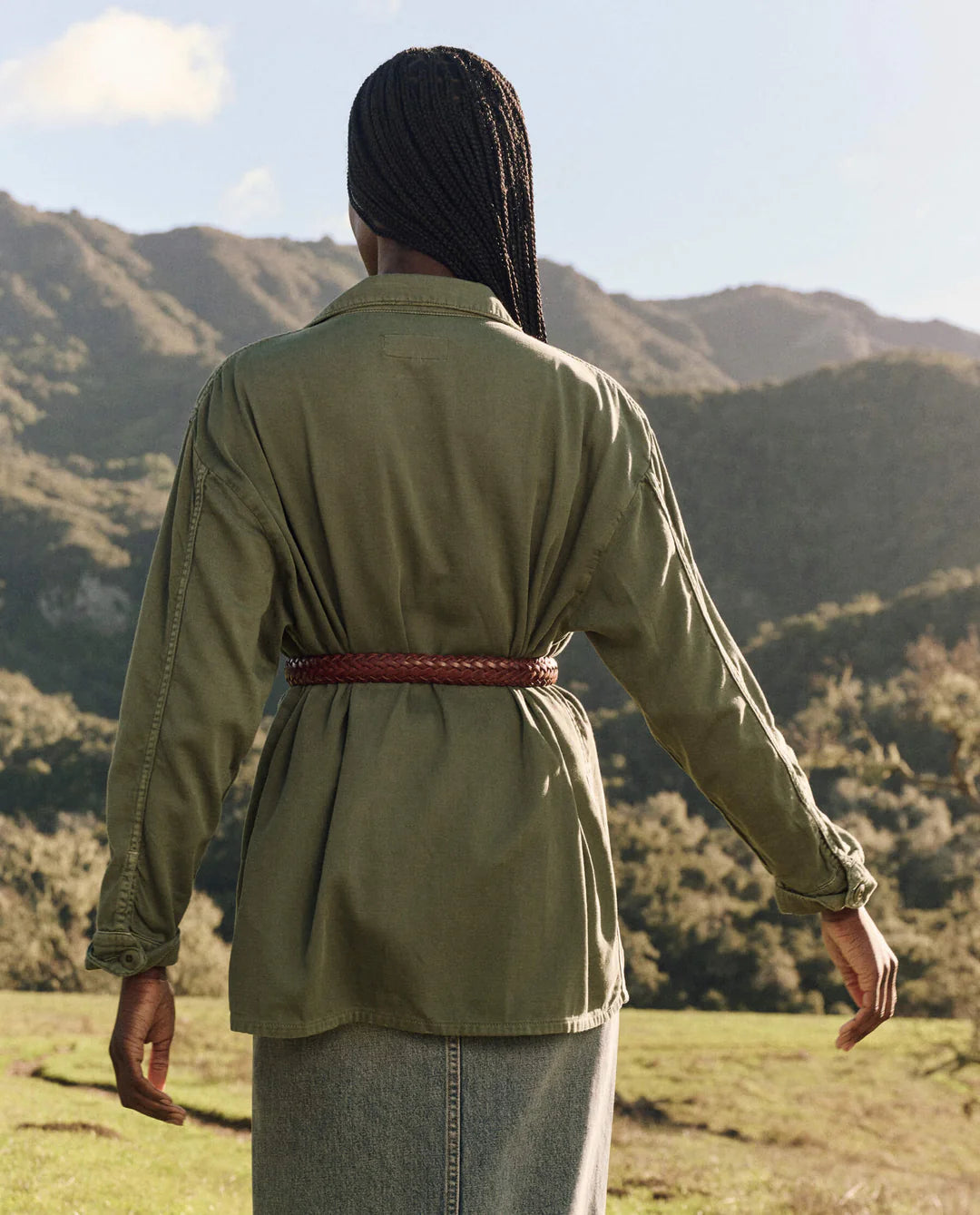 Seen from behind, a person with long braids stands outdoors wearing The Vintage Army Jacket by The Great Inc. and a denim skirt, with green hills and a partly cloudy sky in the background.