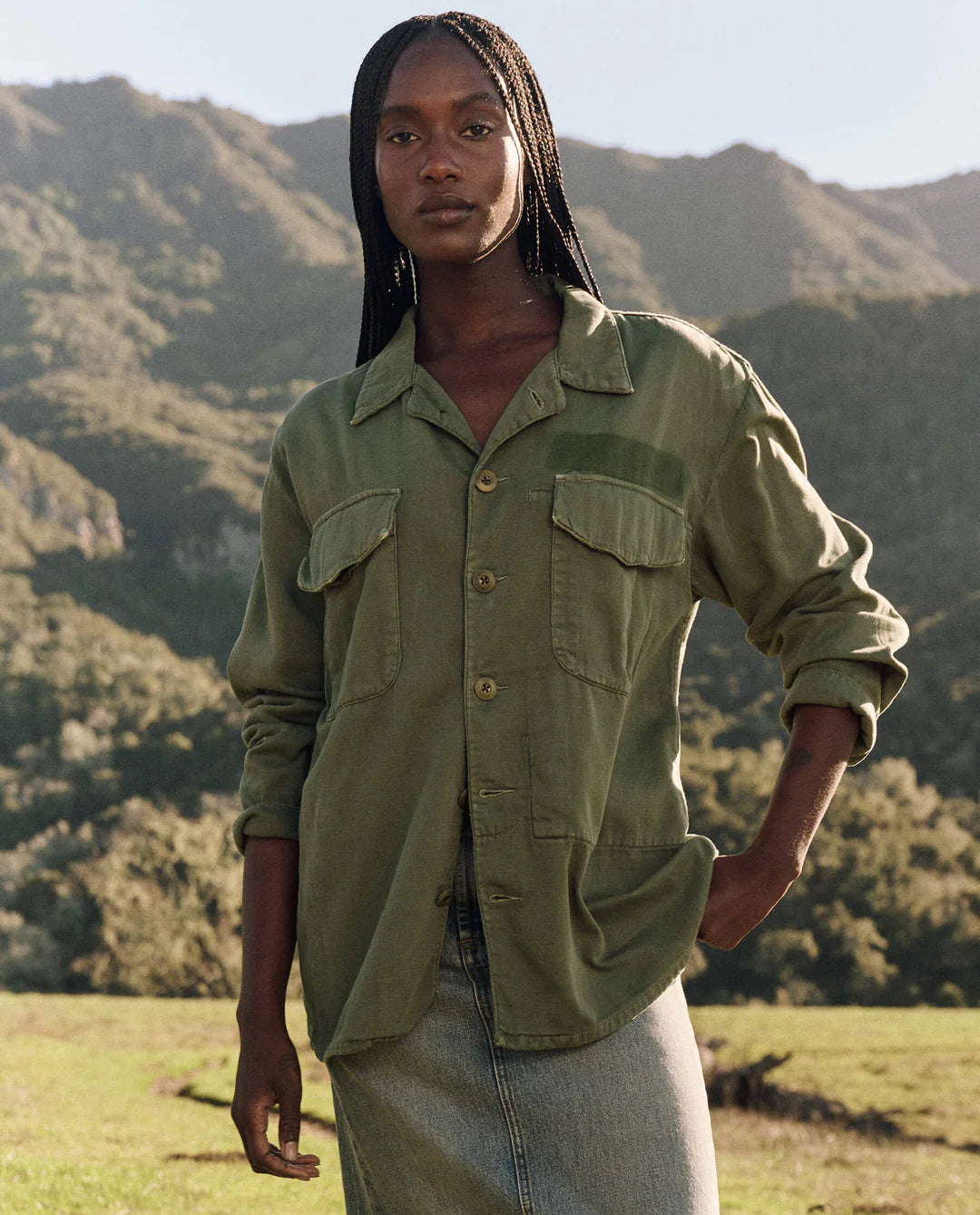 A woman with long braided hair stands outdoors before mountains, confidently wearing The Vintage Army Jacket by The Great Inc. in cotton slub twill and a light denim skirt, one hand in her pocket as she looks at the camera.