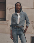 The Great Inc. Howdy Top in denim, styled casually with jeans, set against a rustic beige wall for a relaxed look.