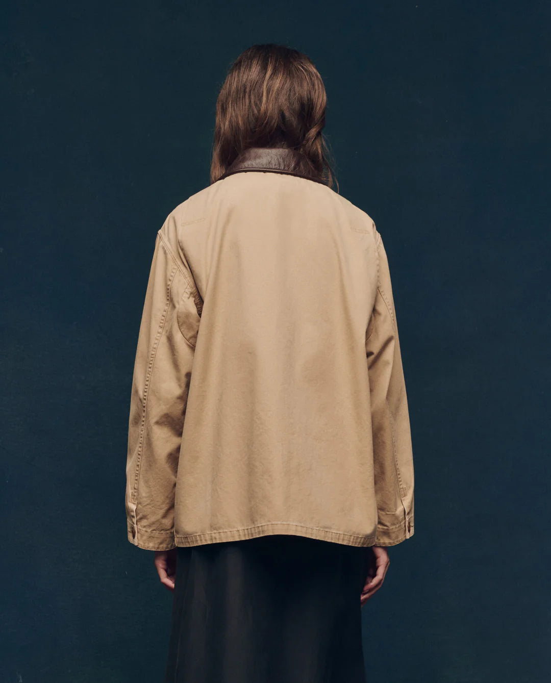A person with long brown hair, wearing The Great Inc.'s The Barn Jacket with utility pockets and a dark skirt, stands facing away from the camera against a dark blue background.