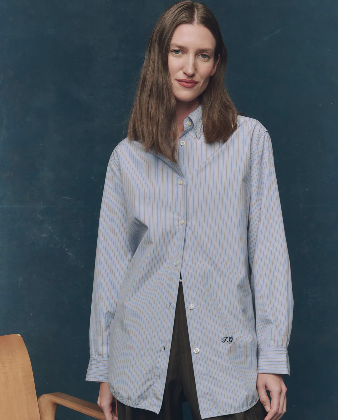 A woman with long brown hair models The Great Inc.'s Menswear Button Up, featuring subtle pinstripes, as she stands before a dark blue background near a wooden chair, smiling gently at the camera.
