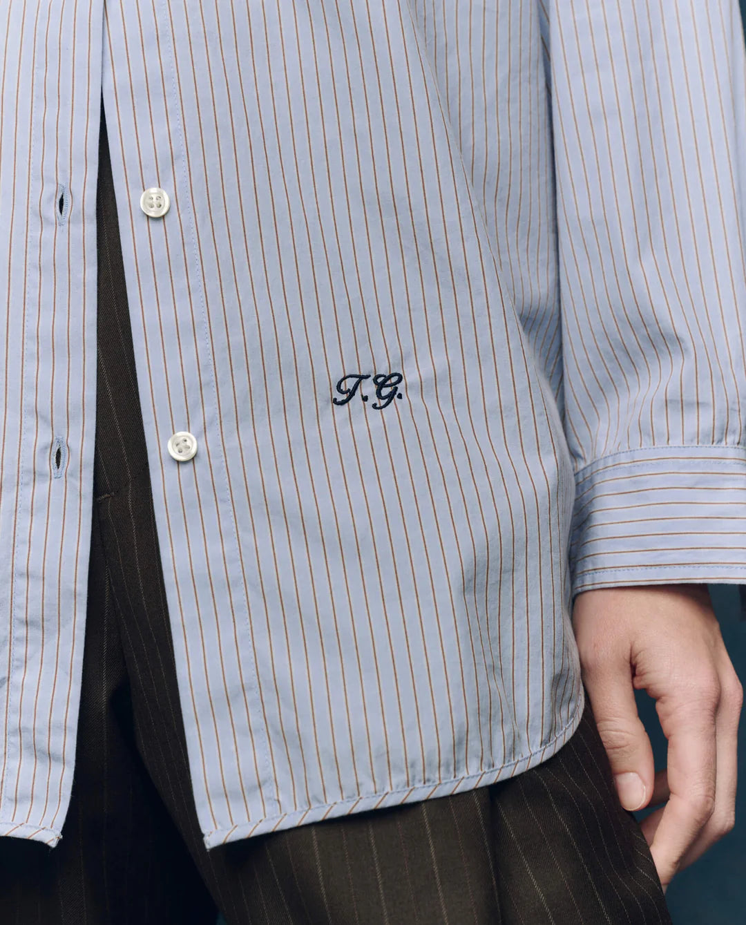 A close-up showcases The Menswear Button Up by The Great Inc., featuring blue and white stripes with “T.G.” embroidered near the hem, styled with classic pinstripe dark pants for a timeless look.