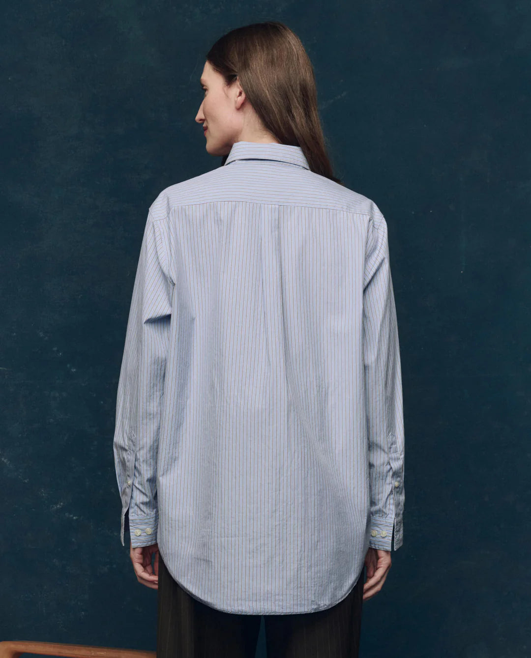 A person with long brown hair, seen from behind, wears The Great Inc.'s The Menswear Button Up and dark trousers, standing against a dark blue textured background.