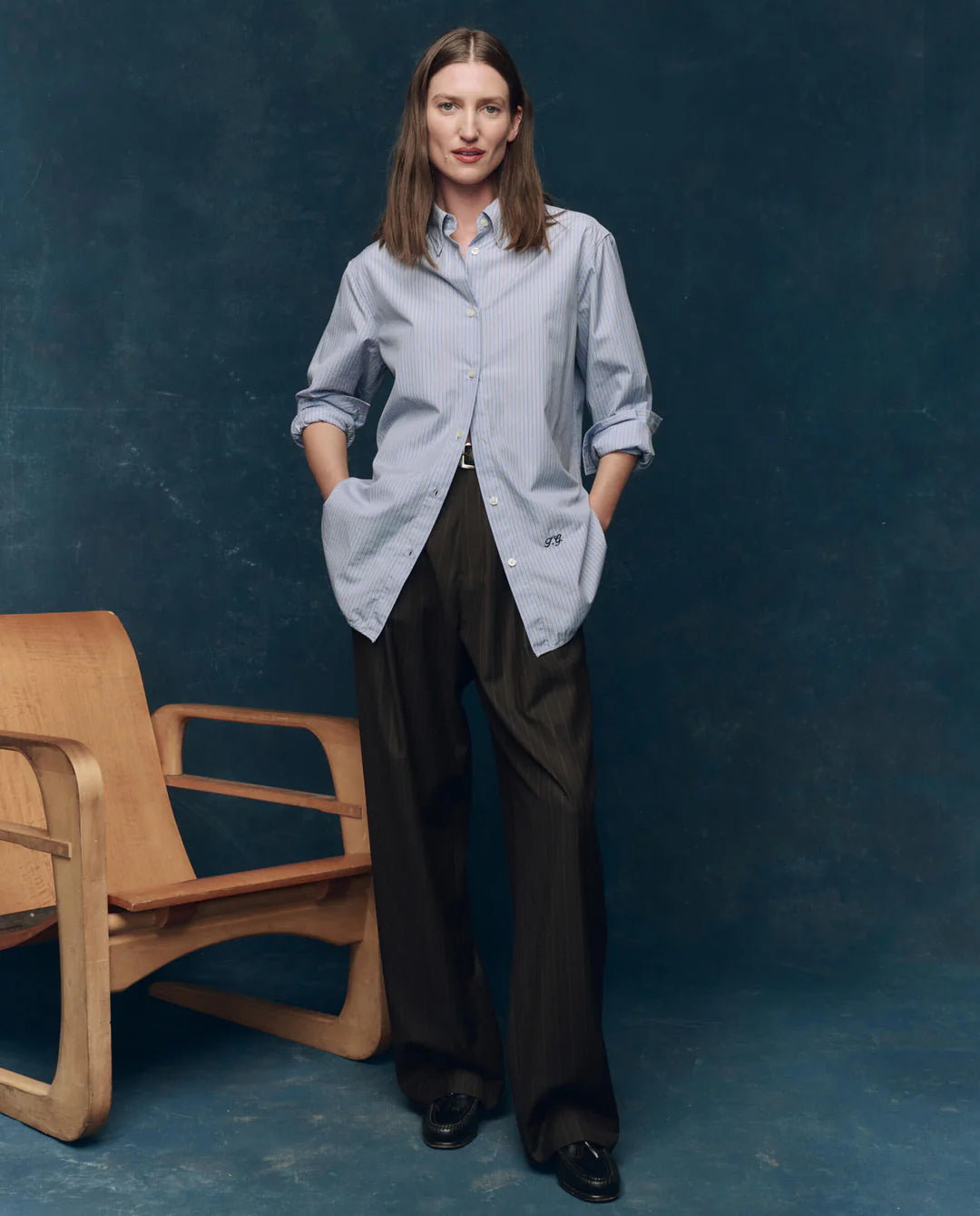 A woman stands against a dark blue background, wearing The Great Inc.’s The Menswear Button Up with rolled sleeves and loose dark trousers, hands in her pockets beside a wooden chair.