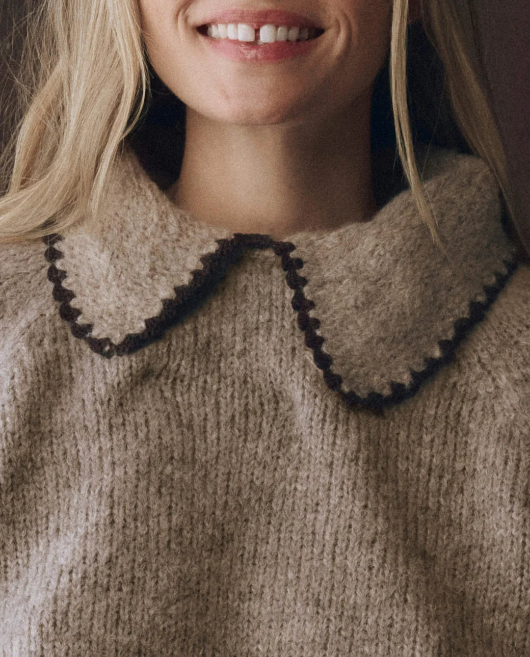 A woman with long blonde hair smiles, wearing The Great Inc.’s Crochet Collar Pullover—a relaxed fit beige sweater with a large rounded crochet collar and dark brown trim. Only her lower face and upper torso are shown.