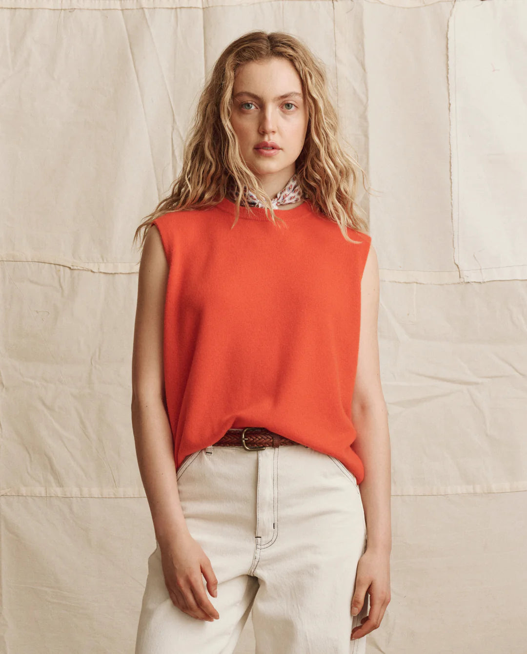 A woman with wavy blonde hair wears The Cashmere Pullover Vest by The Great Inc. in bright orange and high-waisted light beige jeans, standing against a neutral, textured fabric backdrop.