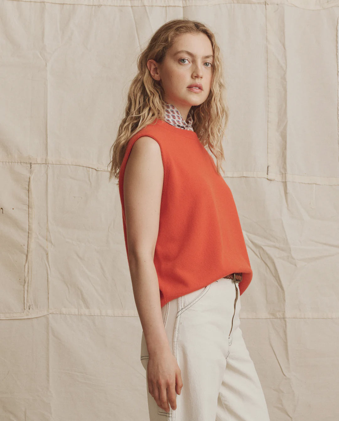 A woman with wavy blonde hair models The Cashmere Pullover Vest by The Great Inc. in bright orange over a patterned collared shirt and white jeans, standing sideways against a textured neutral fabric background.