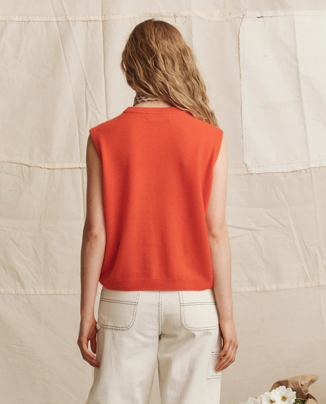 A person with wavy, light brown hair stands with their back to the camera against a cream backdrop, wearing The Great Inc.'s The Cashmere Pullover Vest and high-waisted white pants.