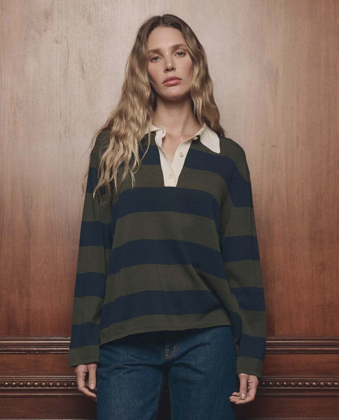 The Academy Polo Sweater by The Great Inc. in a casual style, shown with jeans against a wood panel wall.