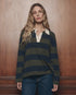 The Academy Polo Sweater by The Great Inc. in a casual style, shown with jeans against a wood panel wall.
