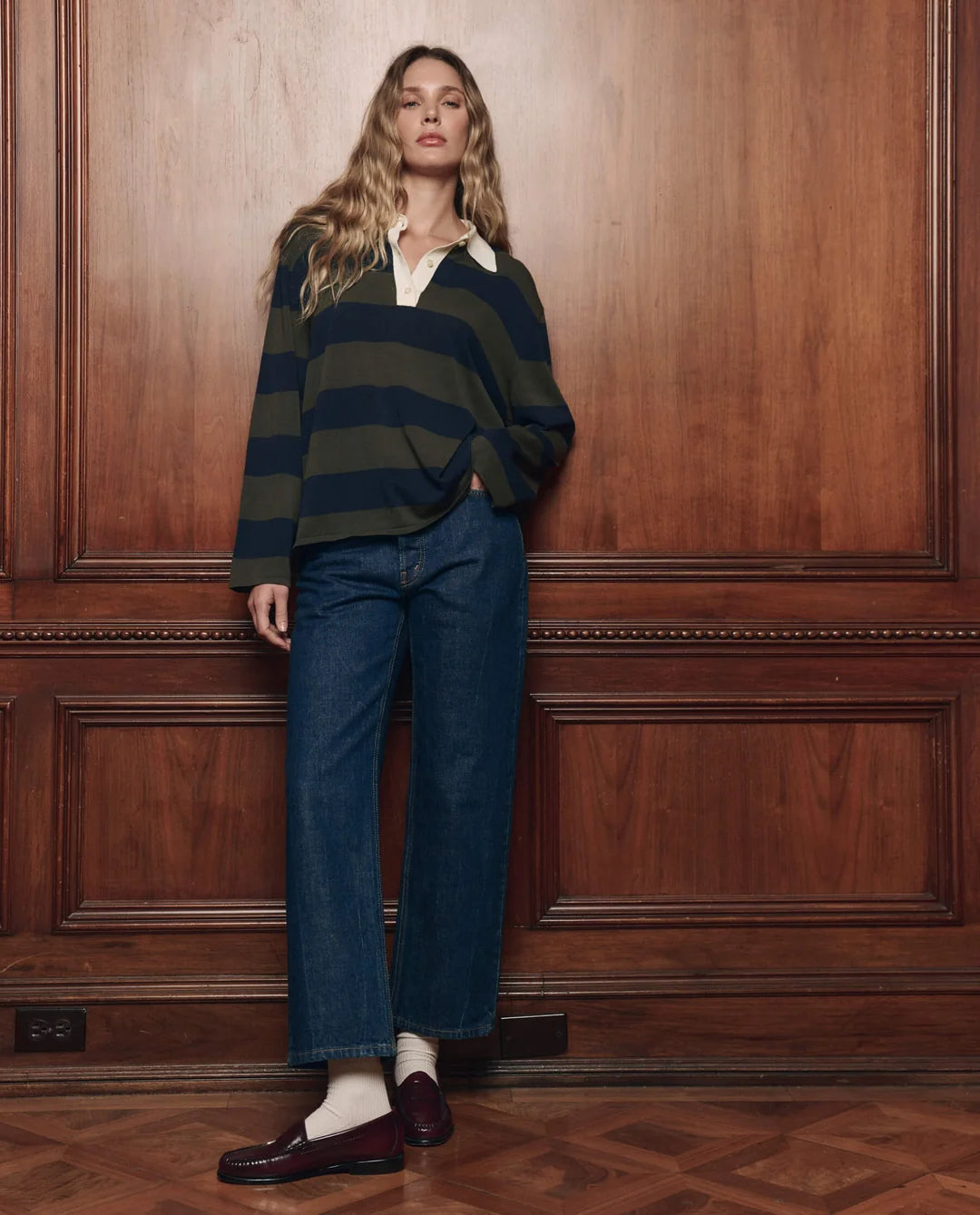 Academy Polo Sweater by The Great Inc. styled with wide-leg jeans and loafers beside wood-paneled wall.