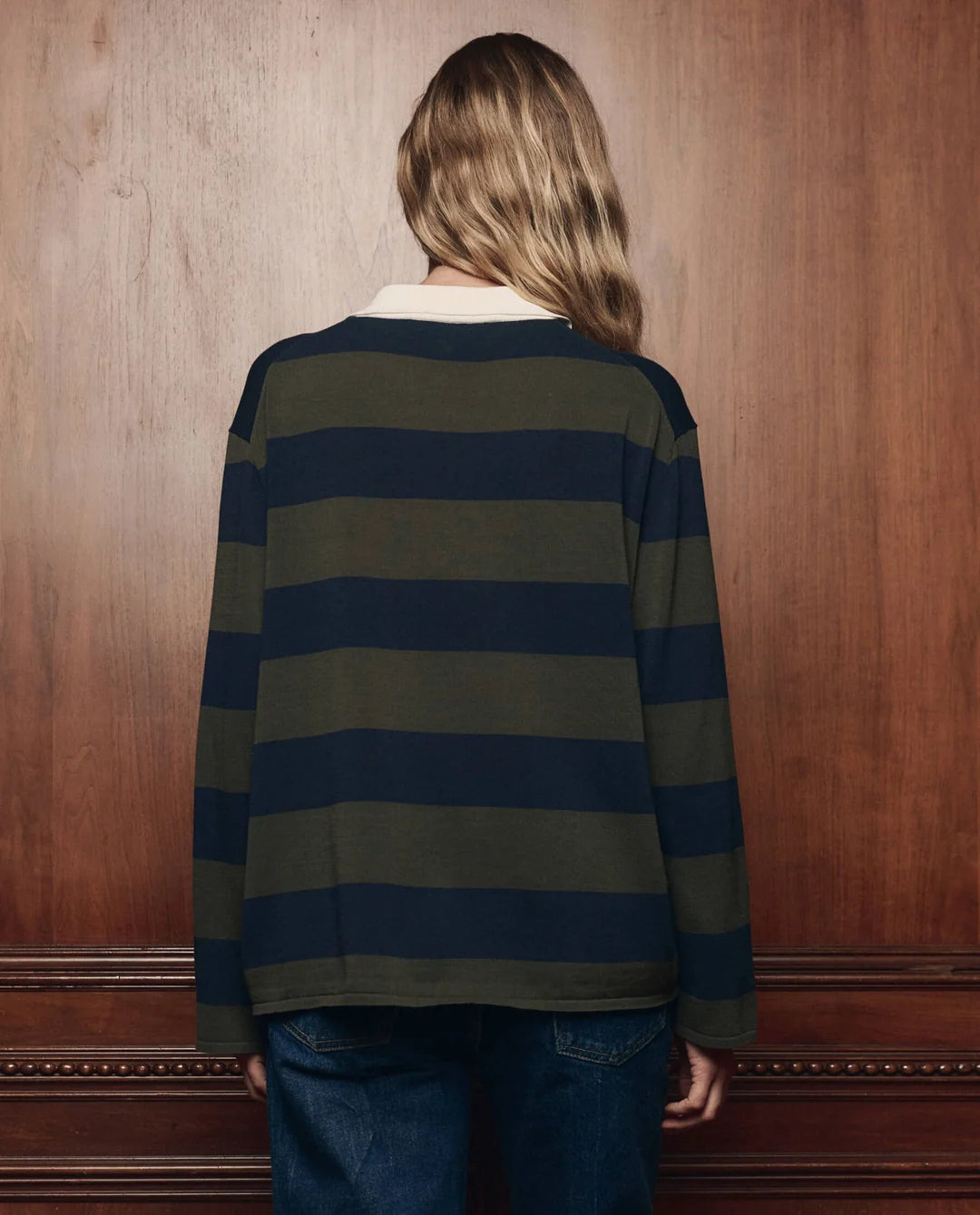 The Great Inc. Academy Polo Sweater in a casual style, worn with jeans near a wooden wall, relaxed setting.