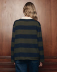 The Great Inc. Academy Polo Sweater in a casual style, worn with jeans near a wooden wall, relaxed setting.
