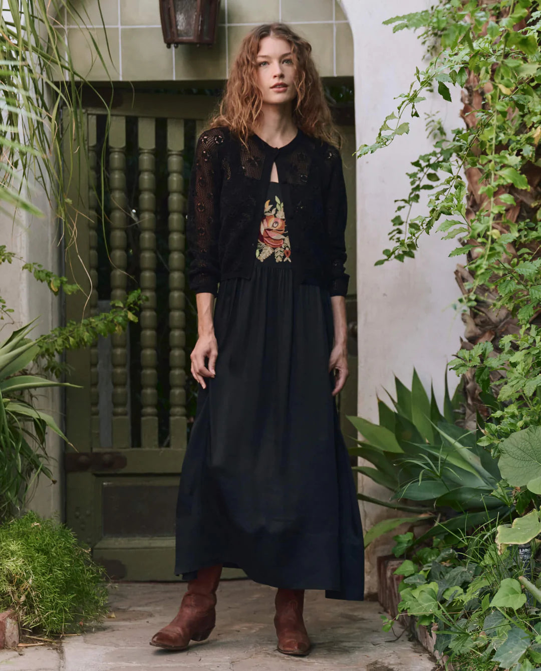 A woman with wavy hair wears The Crochet Cardigan by The Great Inc. over a long black dress and brown boots, standing in a lush garden courtyard with green plants and a decorative wooden door.