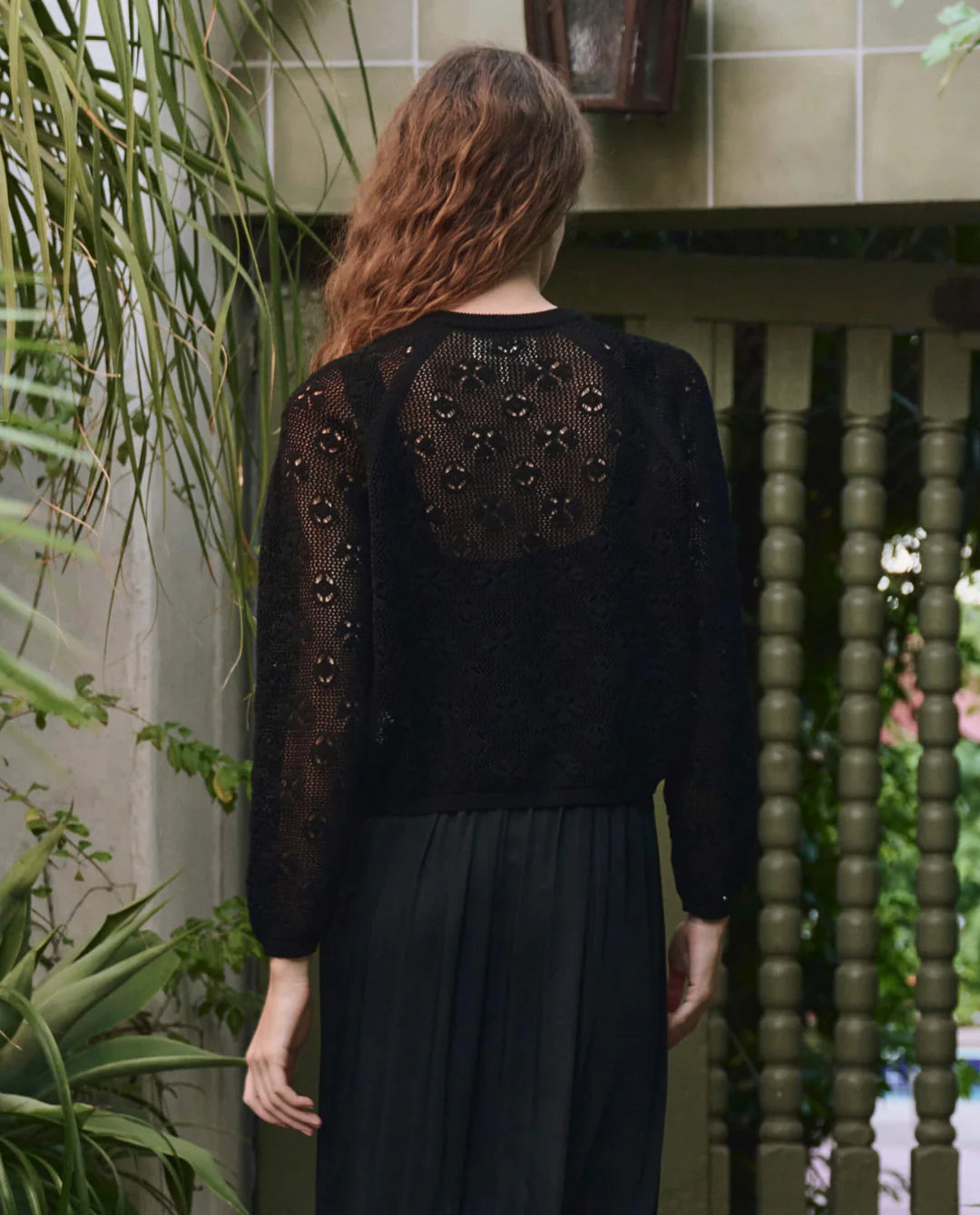 A person with long, wavy hair stands outdoors, facing away from the camera, wearing The Crochet Cardigan by The Great Inc. with a black skirt. They are surrounded by green plants and tiled walls.